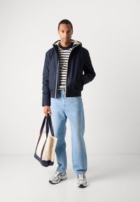 Marineblå bomberjakke med beige foret, stribet skjorte, lyseblå jeans og en beige tote taske med marineblå håndtag. Grå sneakers.