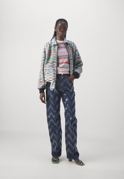 Veste bomber à motifs multicolores, pull blanc à zigzag, et jean large bleu marine avec des paillettes argentées, associés à des sandales à glissière vertes.