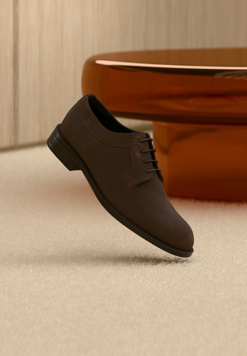 Σκούρο καφέ δερμάτινο παπούτσι για επίσημες εμφανίσεις με κορδόνια, αιωρούμενο πάνω από ένα ανοιχτό μπεζ χαλί με ένα γυαλιστερό τραπέζι κεχριμπαρένιου χρώματος στο φόντο.