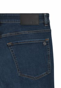 Donkerblauwe denim jeans hebben een achterzak met contrasterende stiksels, een zwart leren merkembleem en een klein metalen logodetail.
