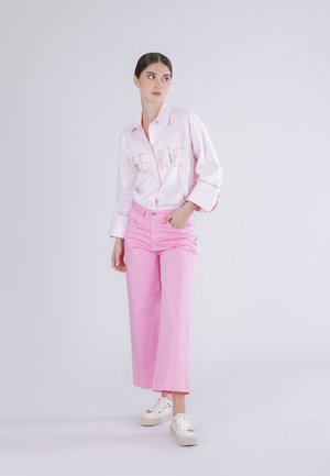 Kvinde stående iført en lyserød skjorte med knapper og pyntet tekst samt høj talje, vidde ben bukser i pink og hvide sneakers.