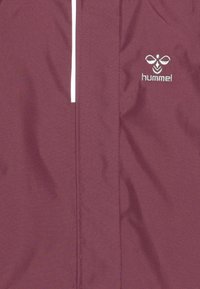 Chaqueta marrón de tela suave con un acento de cremallera blanca vertical y un sutil logo en la parte superior izquierda.