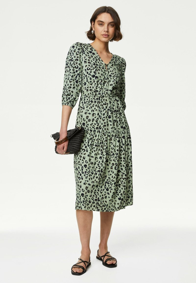 Groen luipaardprint jurkje met driekwart mouwen, V-hals, geplooide taille en gelaagde rok. Gepaarde met een zwarte geweven clutch en sandalen.