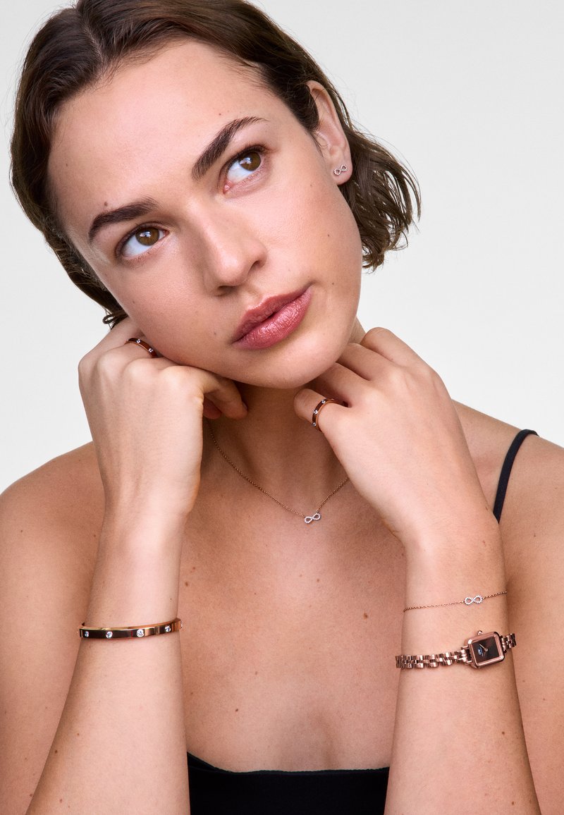 Accessoires en rose gold sur un modèle : un collier superposé avec un symbole de l'infini, un bracelet fin orné de pierres, et une montre à cadran carré.