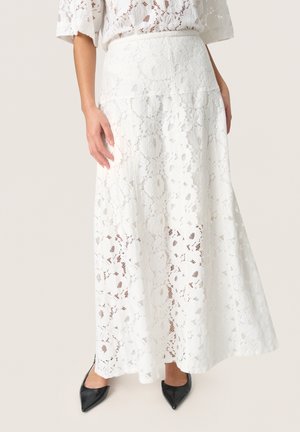 Jupe maxi en dentelle blanche avec un motif floral, texture détaillée, taille ajustée et design fluide. Des escarpins noirs à talons pointus complètent le look.