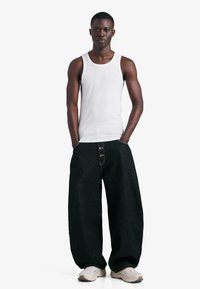 Črni široki kavbojke iz jeansa s kontrastnim šivanjem, kombinirane z belim topom brez rokavov. Model nosi superge nevtralne barve.