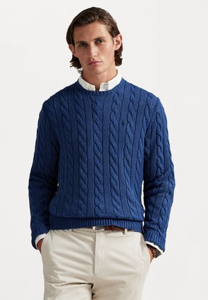 Blå kabelstrikket sweater lavet af et tekstureret materiale, med rund halsudskæring og ribbede manchetter og kant, parret med lyse beige bukser.