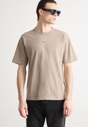 Brun bomulls T-skjorte med kort erm og rund hals, med subtil merkevare på brystet. Brukt med svarte denimbukser.
