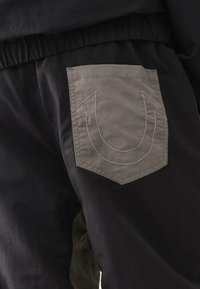 Schwarze Jogginghosen mit einem elastischen Bund, ausgestattet mit einer großen grauen aufgesetzten Tasche auf der Rückseite, die mit kontrastreichem weißen Nähten umrandet ist.