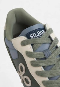 Zapatilla verde y gris que presenta materiales de cuero y textil, punta redonda, cordones gruesos y un acento de logo en el costado con un patrón cosido.