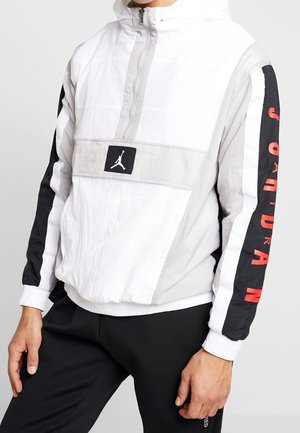 Biała, czarna i szara bluza z kapturem Jordan z przednią kieszenią i czerwonym napisem "JORDAN" na rękawach, noszona z czarnymi spodniami.