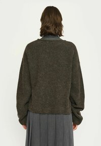 Persona con capelli castani di lunghezza spalle che indossa un maglione largo color marrone scuro e una gonna plissé grigia, girata di spalle contro uno sfondo semplice.