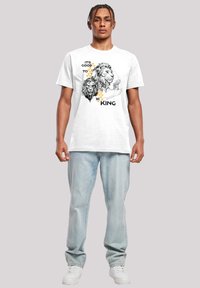 Vit bomull t-shirt med en lejon grafik och text: "DET ÄR BRA ATT VARA KUNG." Modellen bär ljusblå jeans och vita sneakers.