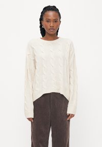 Pulover od pamučne pamučne boje sa uzorkom pletenice, okruglim izrezom i opuštenim krojem. Uparen s tamnosmešnim kord pantalonama.