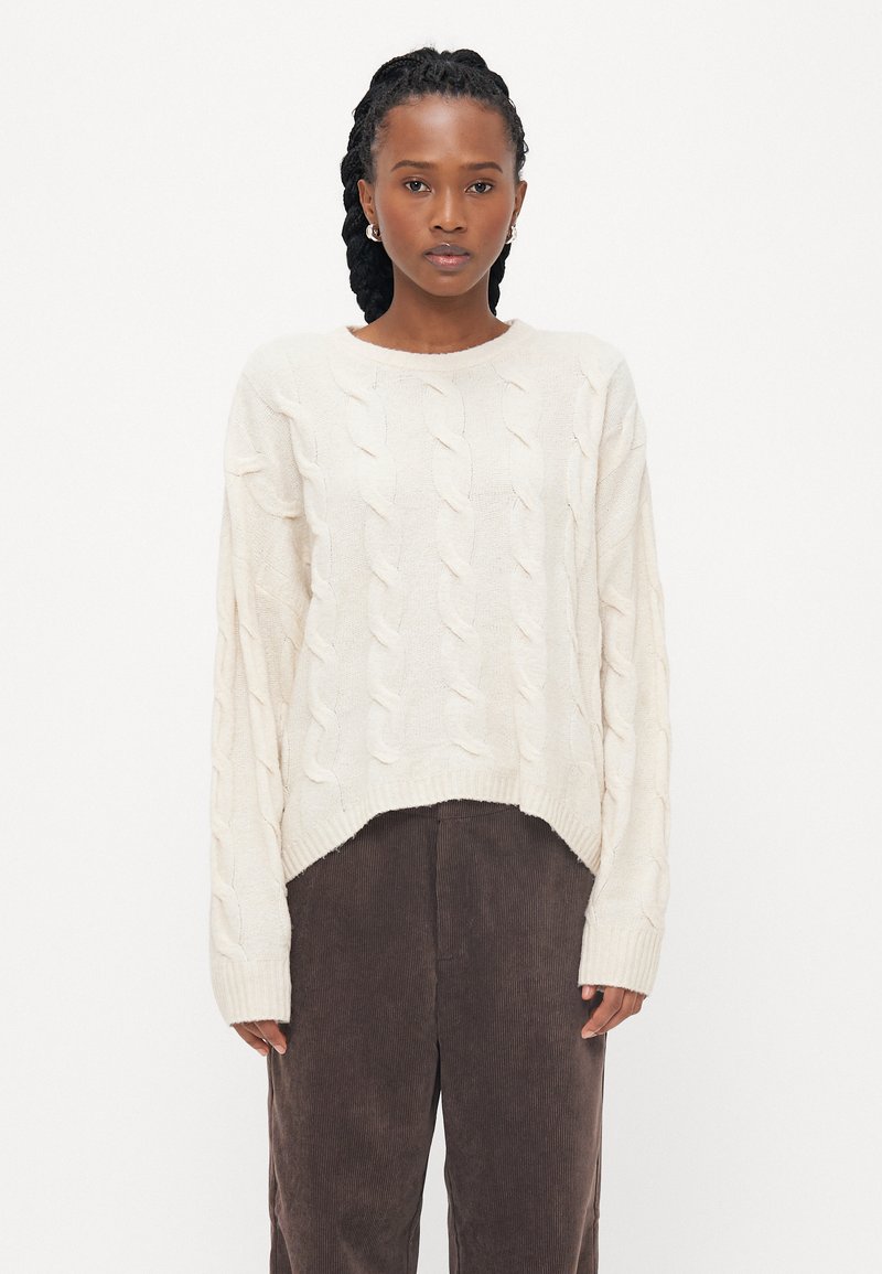 Pulover od pamučne pamučne boje sa uzorkom pletenice, okruglim izrezom i opuštenim krojem. Uparen s tamnosmešnim kord pantalonama.