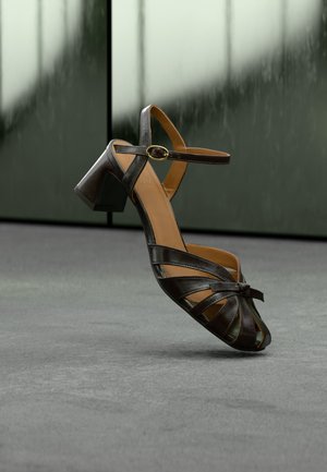 Melnas ādas augstpapēžu sandalēs ar potītes siksnu, sprādzi un sasietām priekšējām siksnām, novietots uz pelēkas grīdas pret aizmiglotu fonu.