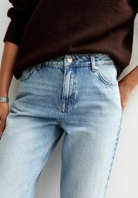 Vaqueros de mezclilla azul claro con una cintura alta, que cuentan con un cierre de botón, diseño de cinco bolsillos y un ligero desvanecimiento. Llevados con un suéter marrón oscuro.