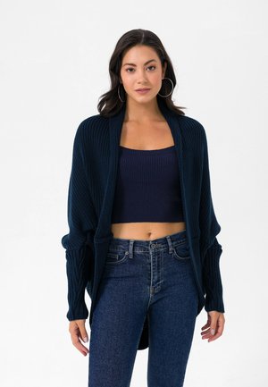 Femme portant un top court bleu marine, un cardigan en maille ouverte assorti, un jean taille haute bleu foncé et de grandes boucles d'oreilles créoles.