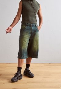 Top sin mangas de color verde con textura de relieve, pantalones cortos de mezclilla de corte relajado que transicionan de verde oscuro a verde claro, y zapatillas deportivas negras con textura y suelas gruesas.