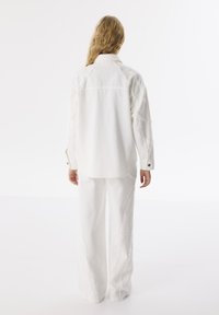 Camicia bianca con bottoni, dettagli cuciti e pantaloni bianchi a gamba larga. Il tessuto sembra testurizzato e informale, adatto per ambienti rilassati.