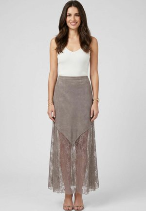 Femme aux longs cheveux bruns portant un haut blanc sans manches et une jupe asymétrique en daim taupe avec une superposition en dentelle et des talons ouverts, debout devant un fond gris.