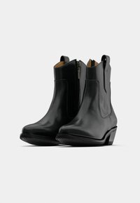 O pereche de cizme negre din piele până la gleznă, cu fermoare laterale, vârfuri rotunde și tocuri joase, pe un fundal alb simplu.