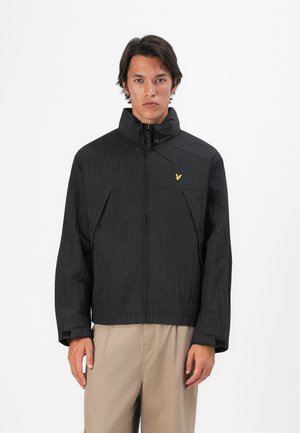 Man wearing a black zip-up jacket with high collar and yellow bird logo, paired with beige pants, standing against a white background.