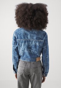 Blue denim jacket with palm tree pattern, cropped design, visible stitching, and branded leather patch. Paired with gray jeans.