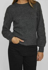 Pull gris en maille texturée avec un col rond et des manches longues légèrement bouffantes. Associé à un pantalon noir, les mains posées dans les poches.