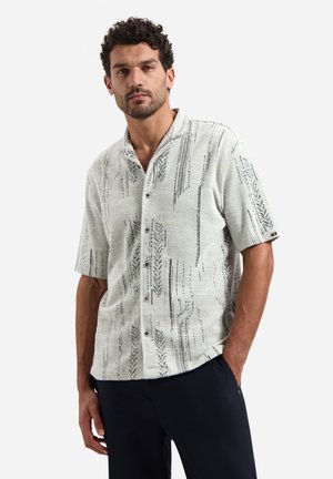 Mann mit lockigem Haar, der ein kurzärmeliges weißes Hemd mit schwarzen vertikalen Mustern trägt, steht mit einer Hand in der Tasche vor weißem Hintergrund.