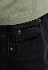 Vaqueros de mezclilla negra con vista de cerca, con herrajes de tono cobre, detalles cosidos y una tela ligeramente texturizada. Camiseta verde visible en la parte superior.