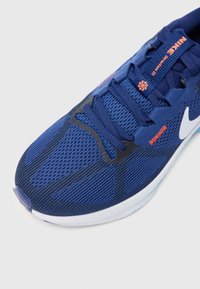 Modra atletska čevlja z teksturirano mrežasto zgornjim delom, črnimi poudarki in belim Nike znakom. vezi so modre z oranžnimi podrobnostmi blagovne znamke.