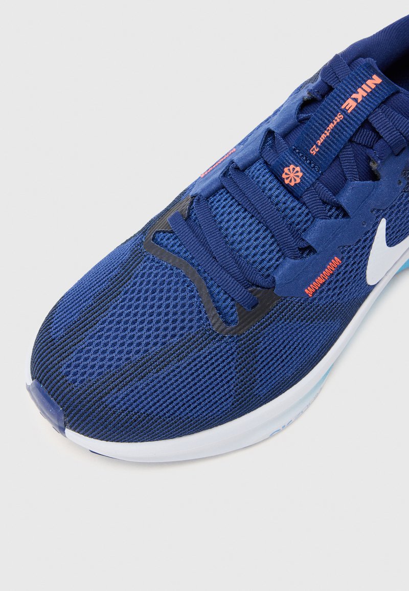 Zapatilla deportiva azul con una parte superior de malla texturizada, acentos en negro y un swoosh blanco de Nike. Los cordones son azules con detalles de marca en naranja.