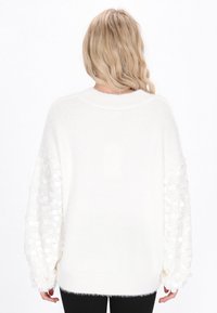 Weißer Strickpullover mit flauschiger Textur, oversized Körper und langen Ärmeln, die mit einem schimmernden kreisförmigen Muster verziert sind.