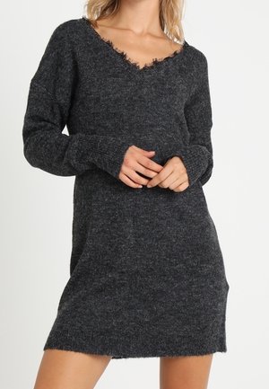 Femme portant une robe-pull en tricot gris foncé à manches longues avec un col en V effiloché, debout devant un fond clair uni.