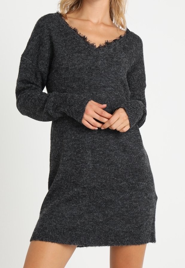 Femme portant une robe-pull en tricot gris foncé à manches longues avec un col en V effiloché, debout devant un fond clair uni.