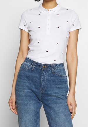 Femme portant un polo blanc à manches courtes avec de petits logos et un jean bleu taille haute, debout devant un fond uni.