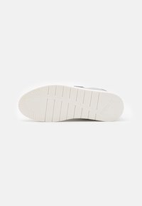 Semelle de sneaker blanche en caoutchouc texturé, avec des rainures horizontales et une légère courbure. Nom de la marque embossé près du talon.