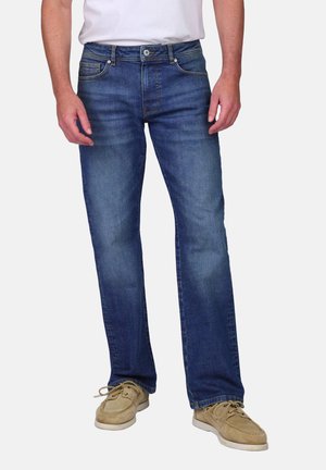 Homme portant un jean en denim bleu, un t-shirt blanc et des chaussures décontractées beige, debout devant un fond clair et uni.