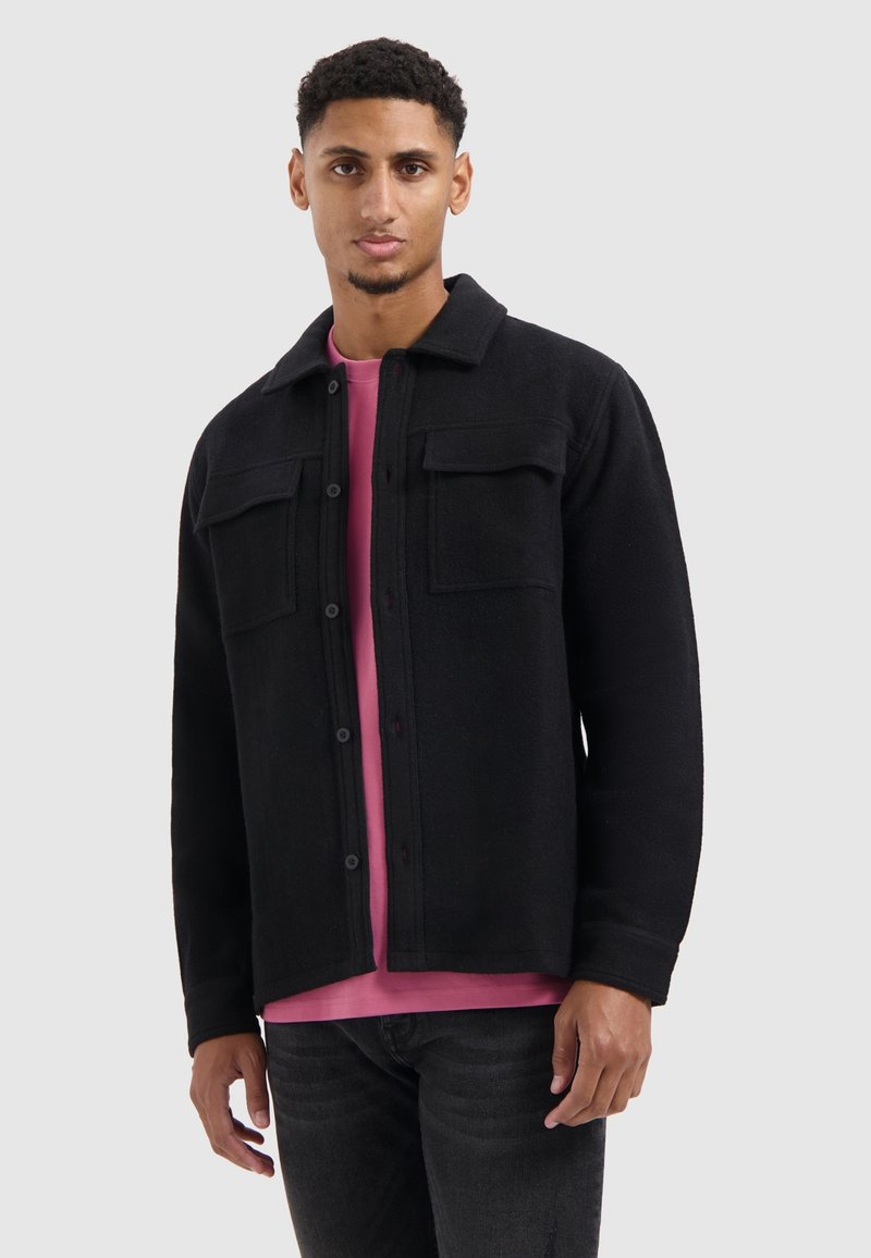 Zwarte fleece jas met kraag, twee borstzakken, knoopsluiting en een relaxte pasvorm, gedragen over een roze shirt en donkere jeans.