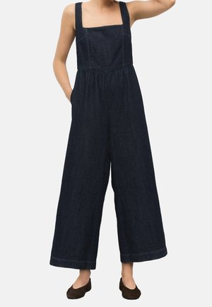 Femme portant une combinaison en jean bleu foncé à jambes larges, encolure carrée et bretelles épaisses, associée à des chaussures plates marron foncé.