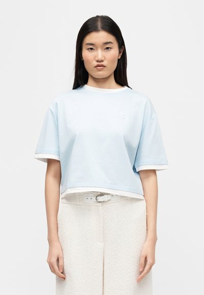 Γυναίκα με μακριά σκούρα μαλλιά που φοράει γαλάζιο κοντό μπλουζάκι πάνω από λευκό εσώρουχο και λευκό υφασμάτινο παντελόνι με ζώνη.