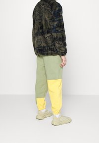 Flauschige grüne und marineblaue gemusterte Jacke mit hohem Kragen; hellgrüne und gelbe Blockfarben-Sweatpants; beigere Sneakers.