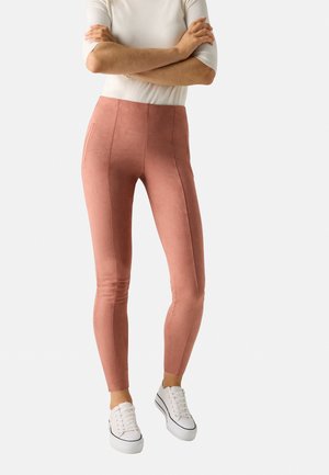 Leggings de ante rosa de cintura alta, con bolsillos laterales y un corte tapered, combinados con zapatillas blancas y una blusa ajustada.