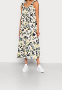 Robe maxi florale en crème avec des motifs verts, bleus et jaunes, dotée de bretelles froncées et d'une silhouette fluide, associée à des baskets blanches.