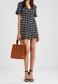 Femme portant une robe noire avec des cœurs blancs, tenant un sac à main marron et portant des sandales à talons hauts noires avec bride à la cheville.