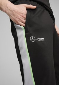 Černé sportovní kalhoty mají kapsu, šedé boční panely s zelenými akcenty a potištěné logo v bílé barvě na spodní části nohavice.