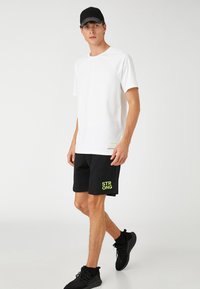 Camiseta atlética blanca de manga corta, combinada con pantalones cortos negros que presentan un estampado neón de "FUERTE". Zapatillas negras completan el conjunto.