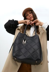 Eine Frau hält eine große schwarze gesteppte Handtasche, trägt einen beigen Mantel und eine braune Mütze, mit einem neutralen Hintergrund, der die Details der Tasche betont.