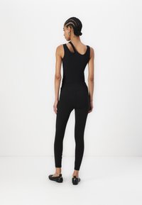 Zwarte sleeveless bodysuit met een ronde rug, gecombineerd met hoge zwarte leggings. Het model draagt zwarte flats, wat de gladde stoftextuur benadrukt.